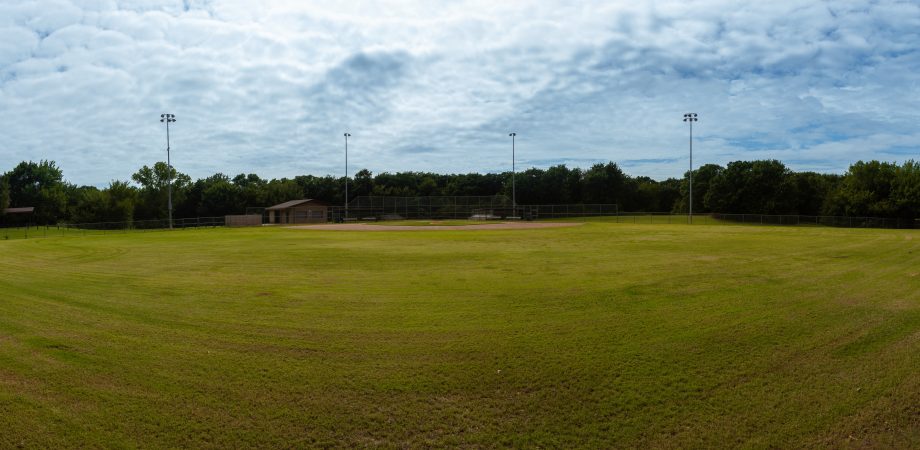 A panoramic view of an empty baseball field in a city park looking toward home base from centerfield on a cloudy, overcast summer day.